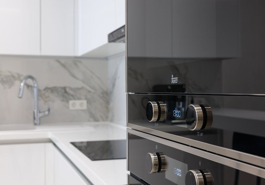 Close-up of a modern built-in oven with a stainless steel finish, featuring control knobs and a digital display showing the time 13:30, situated in a contemporary kitchen. The surroundings include a w
