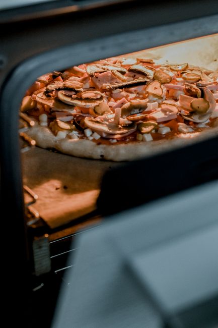 Close-up view of a pizza baking inside an oven with a dark metal interior. The pizza queso base is topped with sliced mushrooms and other ingredients, visible through the oven's glass door. The oven's interior surface appears clean, with a slight glow from the baking process. The oven is part of a residential kitchen setting, and the scene emphasizes surface cleaning and sanitisation in domestic environments. Oven Cleaning Enfield offers professional oven cleaning services to maintain hygienic and safe cooking appliances, ensuring deep cleaning of surfaces like oven walls, racks, and glass doors for optimal performance and hygiene.
