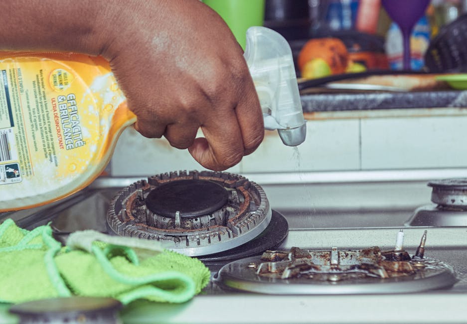 A close-up view of a person's hand spraying a cleaning solution from a yellow bottle labeled 'Eucalyptus & Brulance' onto a gas stove burner in a kitchen. The stove surface is metallic with visible signs of previous use, including rust and grease. The burner is surrounded by a black metal grate, and part of a green cloth is draped nearby. In the background, kitchen items such as colorful objects and containers are visible, indicating a domestic setting. The lighting highlights the stainless steel and ceramic surfaces, emphasizing cleanliness and preparation for deep cleaning. This image reflects surface cleaning and maintenance activities, demonstrating the expertise of Oven Cleaning Enfield in oven and kitchen surface sanitation.