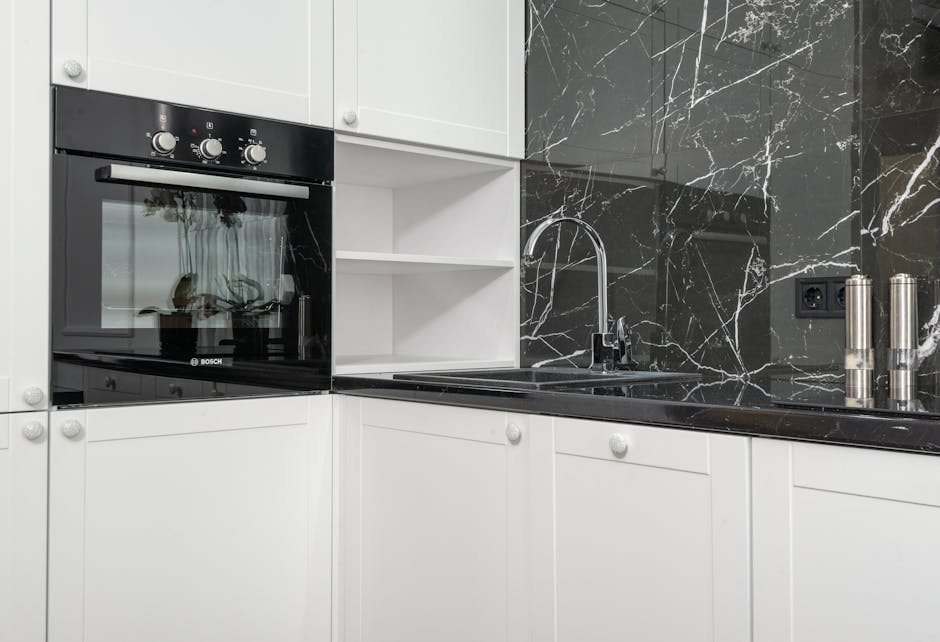 A modern kitchen featuring a built-in Bosch oven with a black glass door and stainless steel control knobs, set within white cabinetry. To the right of the oven, there is a black marble countertop wit